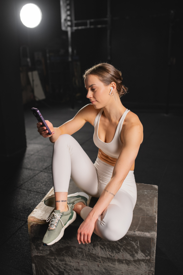 Fit young woman resting after training in gym, scrolling on smartphone, checking workout app. Routine workout for physical and mental health.