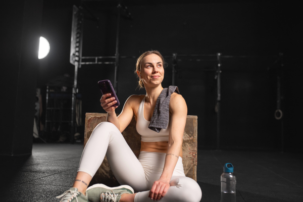 Fit young woman resting after training in gym, scrolling on smartphone, checking workout app. Routine workout for physical and mental health.