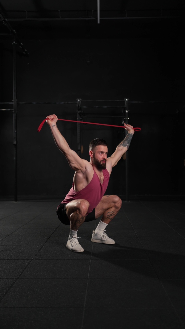 Video of handsome muscular man training with resistance band in gym, squatting. Routine workout for physical and mental health.