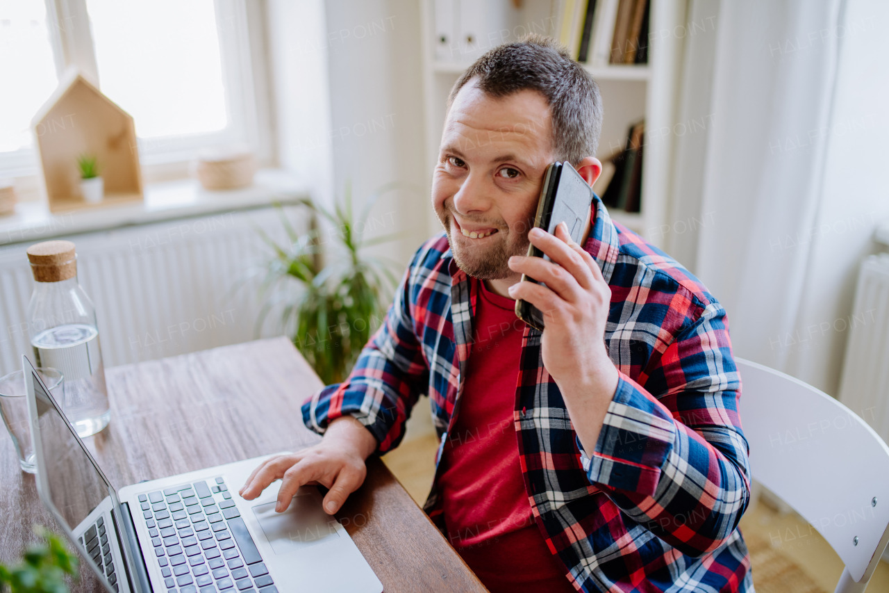 A young man with Down syndrome sitting at desk at home and using smartphone and laptop, looking at camera and smiling. Concept of integration disabled people, home office.