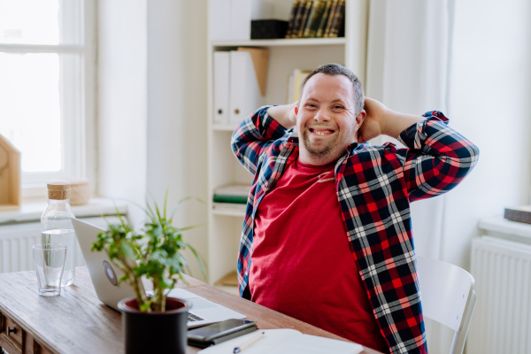 A young man with Down syndrome sitting at desk at home and using laptop, looking at camera and smiling.