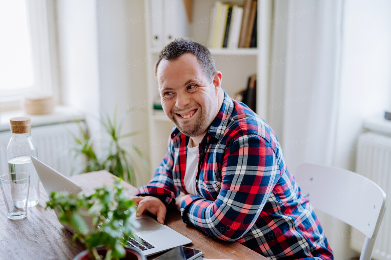 A young man with Down syndrome sitting at desk at home and using laptop, looking at camera and smiling.