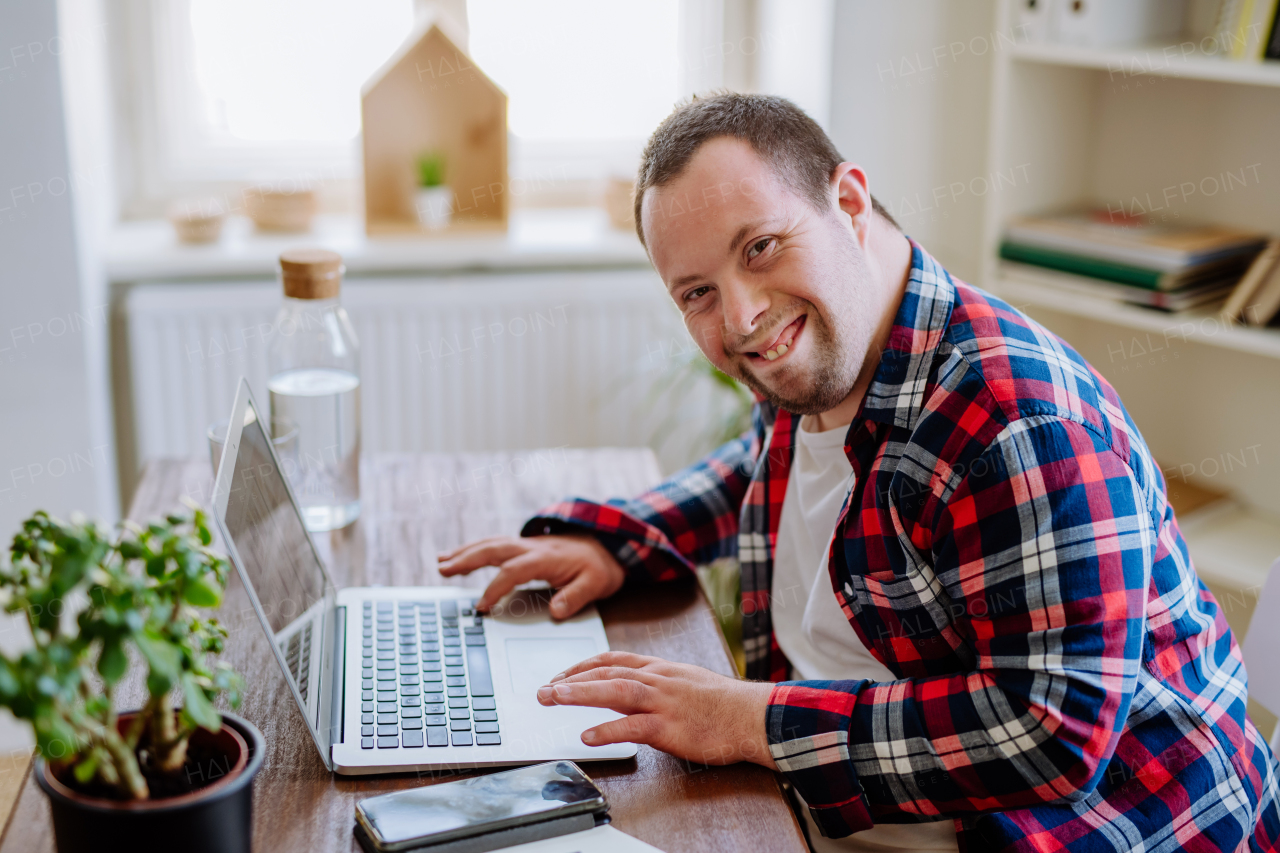 A young man with Down syndrome sitting at desk at home and using laptop, looking at camera and smiling.