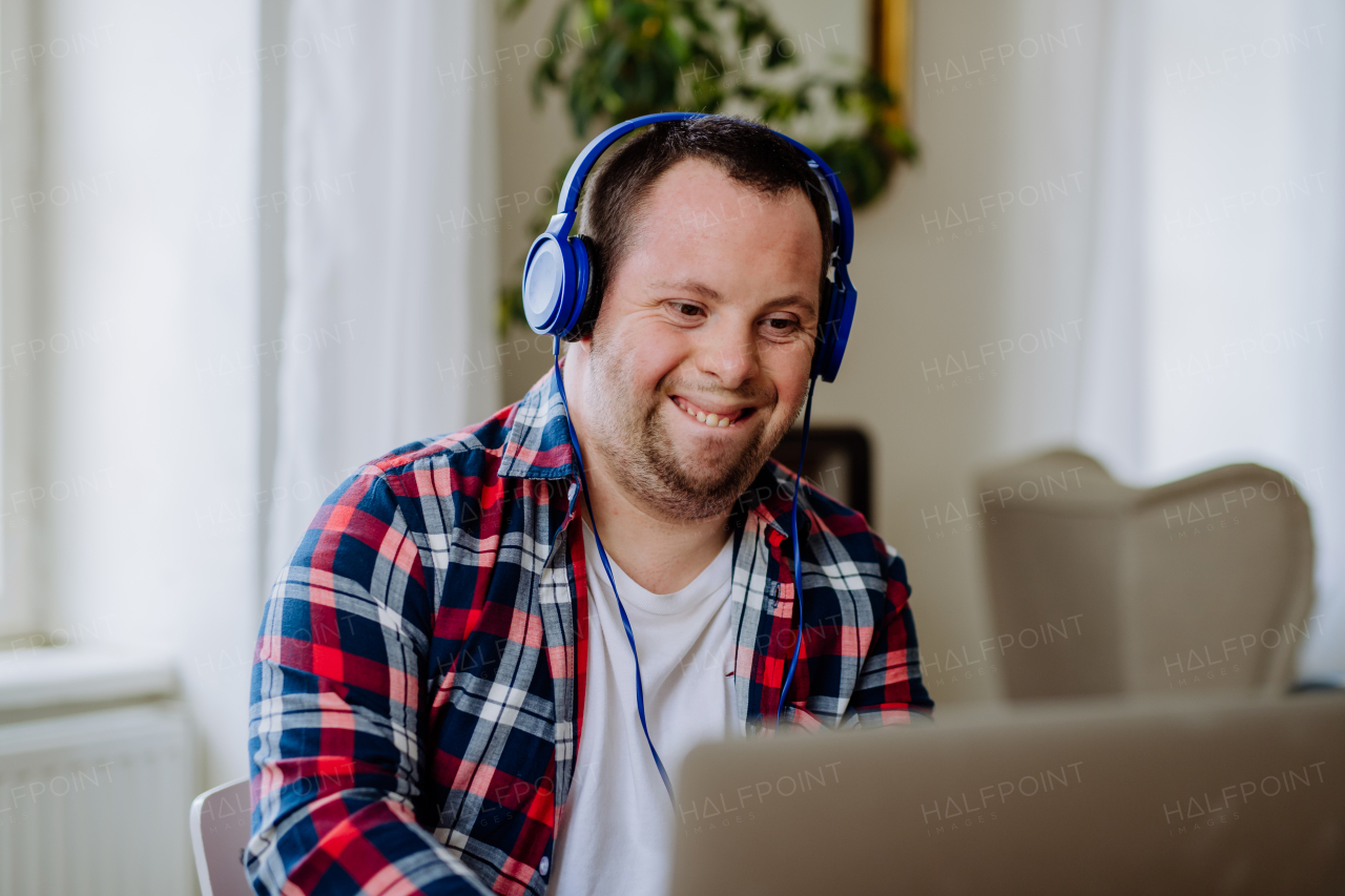 A young man with Down syndrome sitting at desk in office and using laptop, listening to music from headphones.