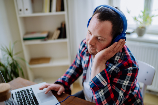 A young man with Down syndrome sitting at desk in office and using laptop, listening to music from headphones.