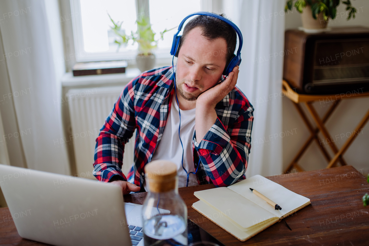 A young man with Down syndrome sitting at desk in office and using laptop, listening to music from headphones.