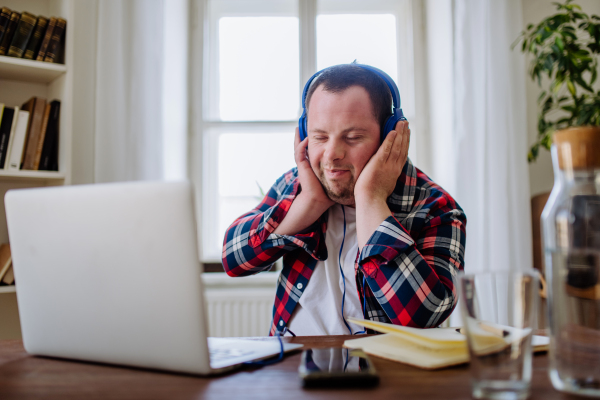 A young man with Down syndrome sitting at desk in office and using laptop, listening to music from headphones.