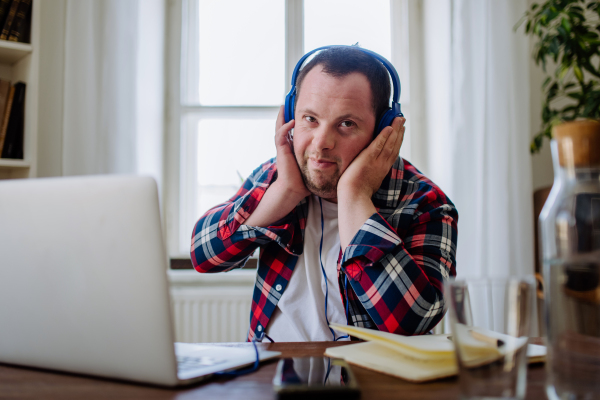 A young man with Down syndrome sitting at desk in office and using laptop, listening to music from headphones.