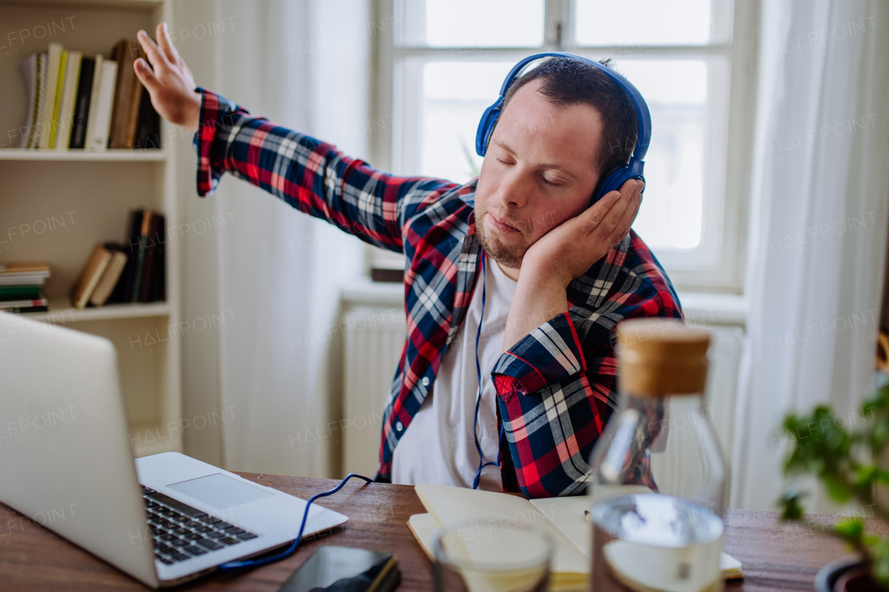 A young man with Down syndrome sitting at desk in office and using laptop, listening to music from headphones.