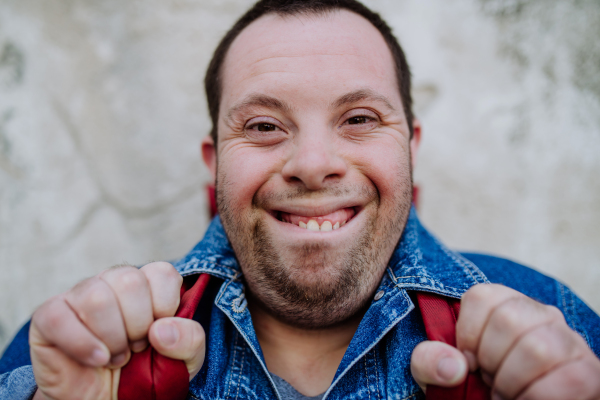 A close-up portrait of happy young man with Down sydrome with backpack in street, smiling.