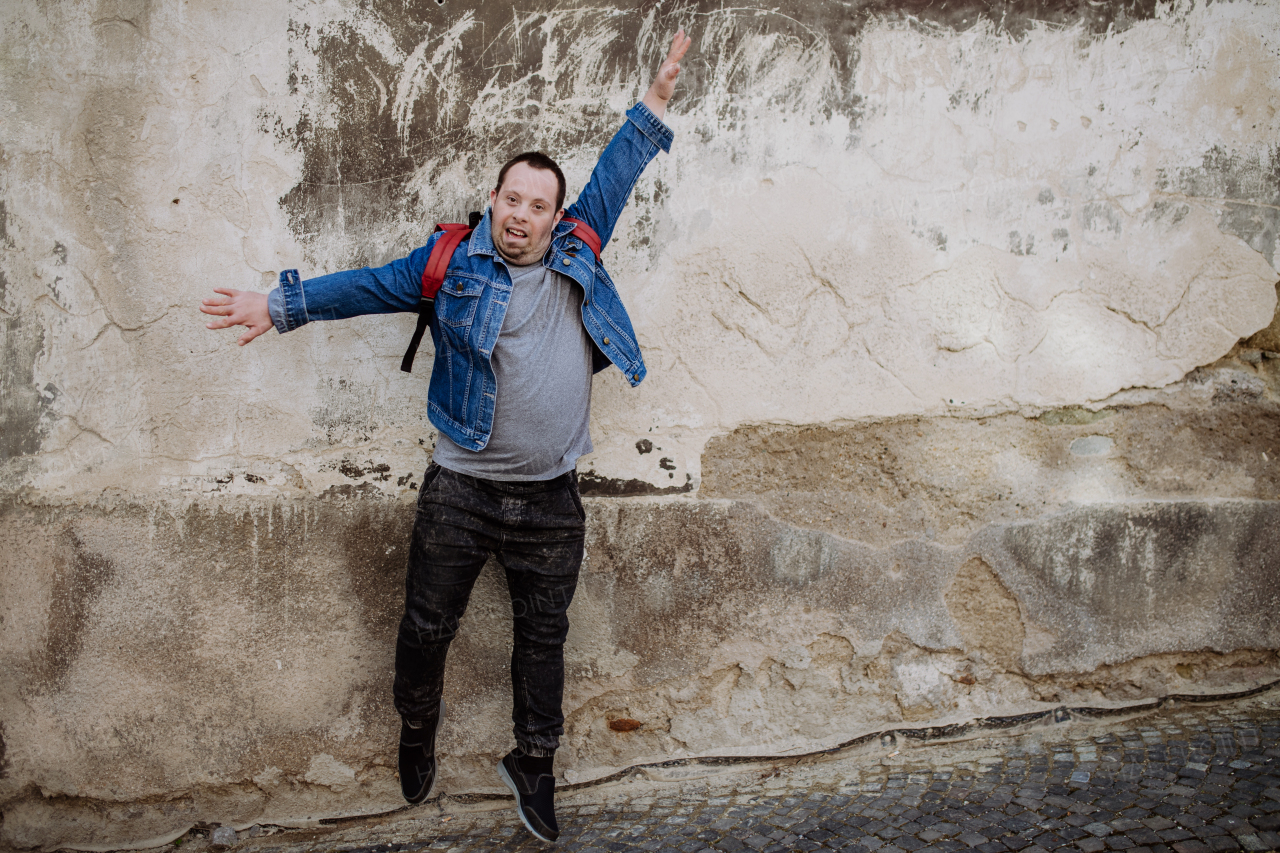 Cheerful young man with Down sydrome jumping in the street, in front of a wall with raising arm, happy gesture.