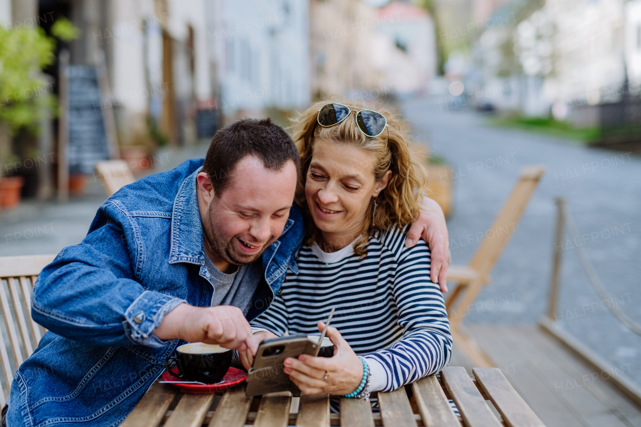 A portrait of happy young man with Down syndrome with his mother sitting in cafe outdoors talking and looking at smartphone.