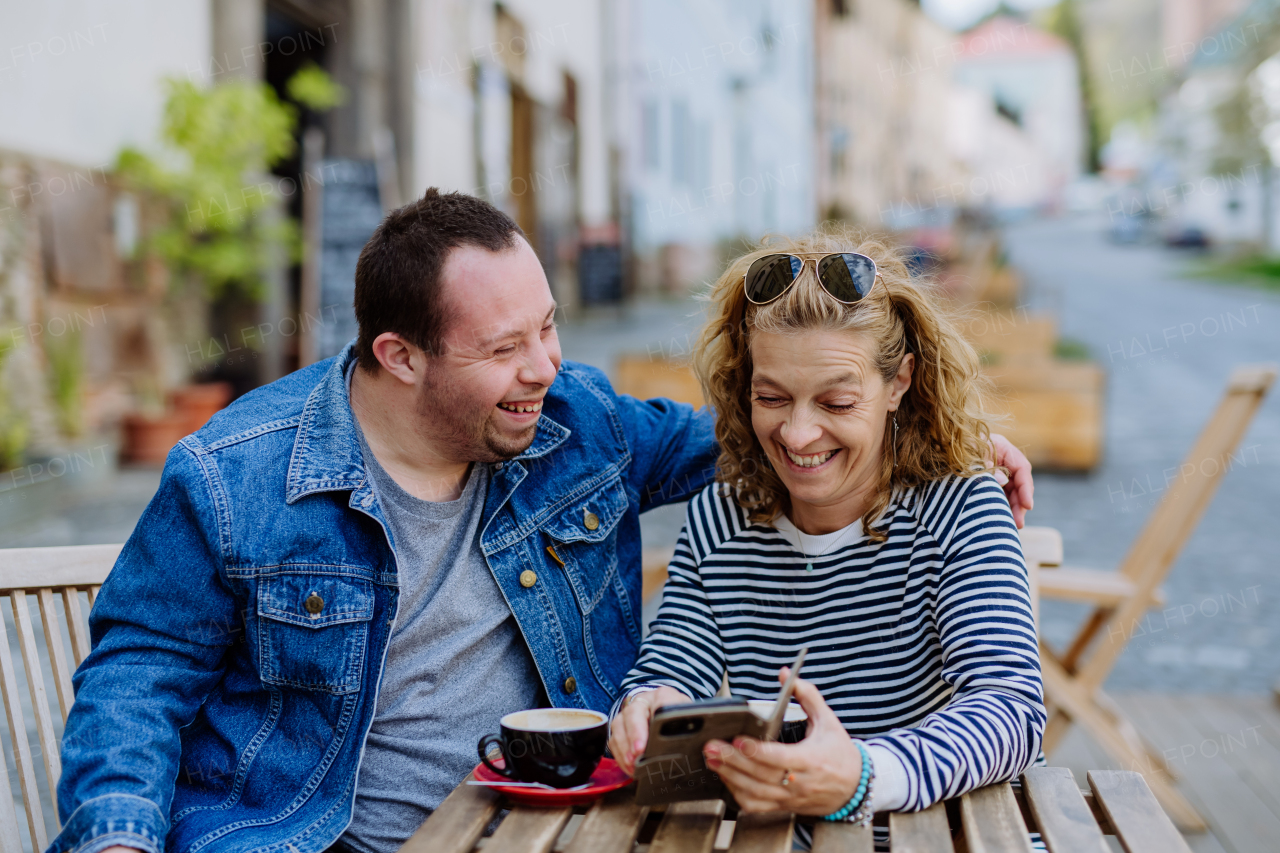 A portrait of happy young man with Down syndrome with his mother sitting in cafe outdoors and talking.