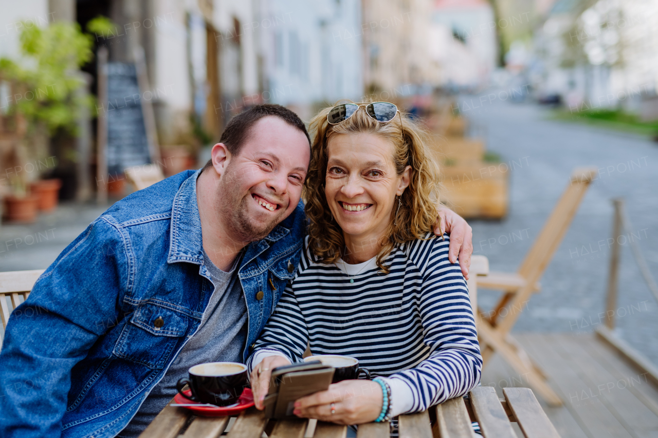 A portrait of happy young man with Down syndrome with his mother sitting in cafe outdoors and talking.