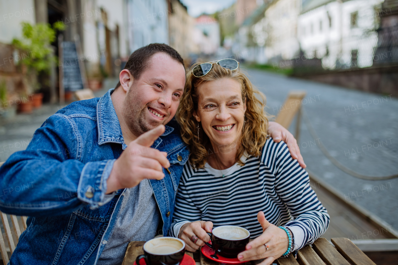 A portrait of happy young man with Down syndrome with his mother sitting in cafe outdoors and talking.