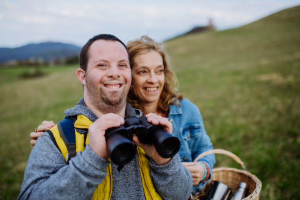 A portrait of happy young man with Down syndrome with his mother resting in nature, sitting and looking trough binoculars.