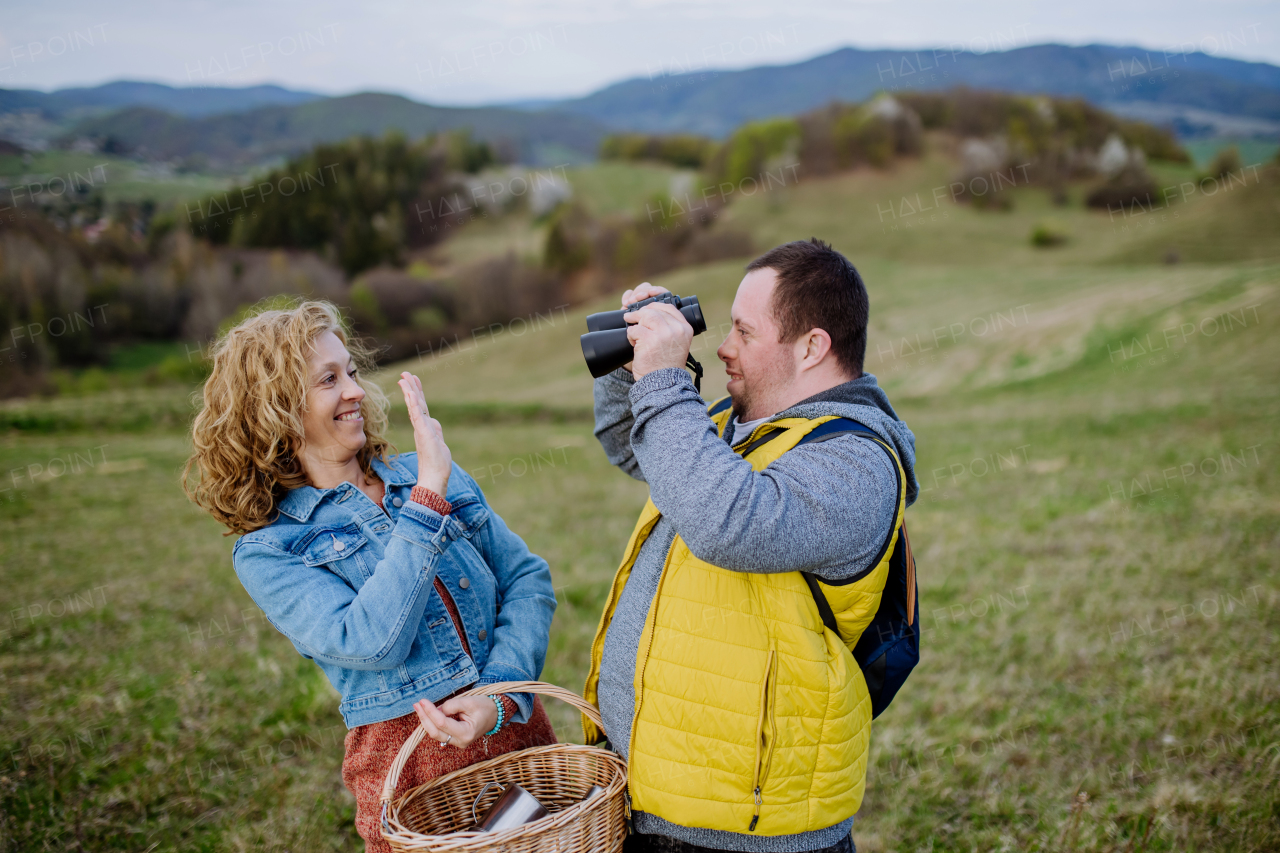 A portrait of happy young man with Down syndrome with his mother resting in nature, sitting and looking at view with binoculars.