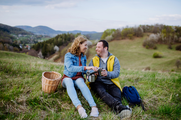 A portrait of happy young man with Down syndrome with his mother resting in nature, sitting and drinking tea.