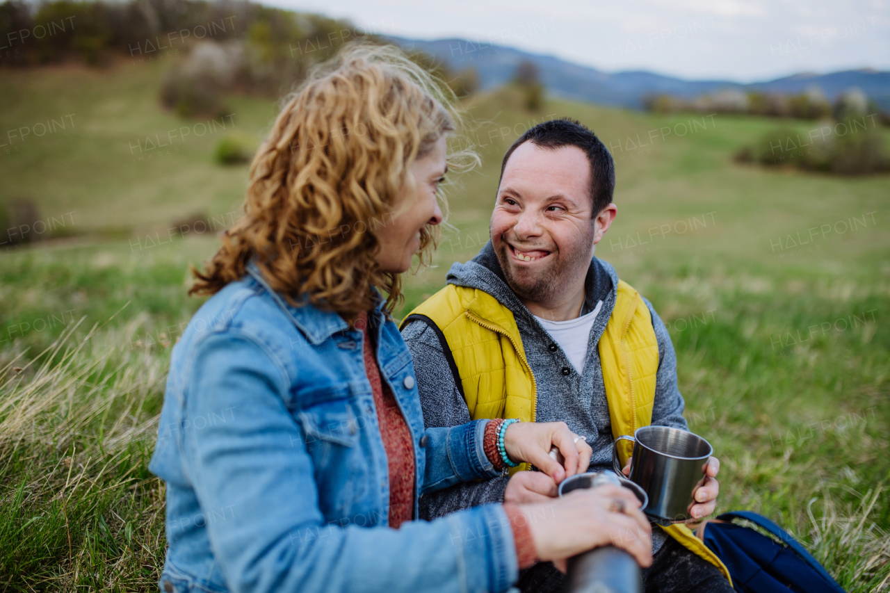 A portrait of happy young man with Down syndrome with his mother resting in nature, sitting and looking at view.