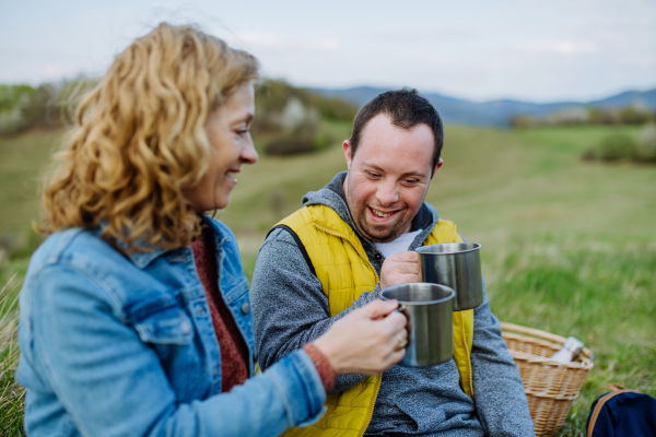 A portrait of happy young man with Down syndrome with his mother resting in nature, sitting and drinking tea.