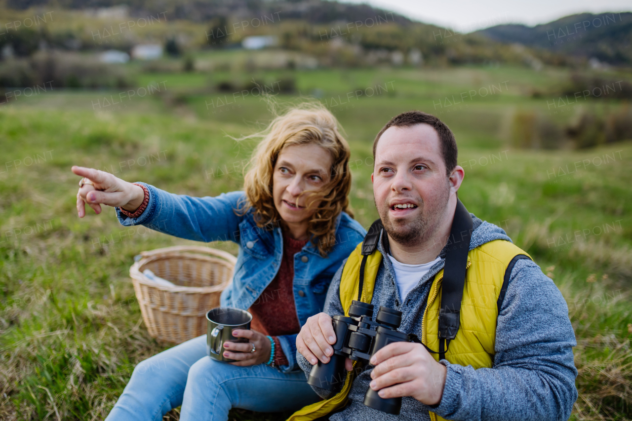 A portrait of happy young man with Down syndrome with his mother resting in nature, sitting and looking at view with binoculars.