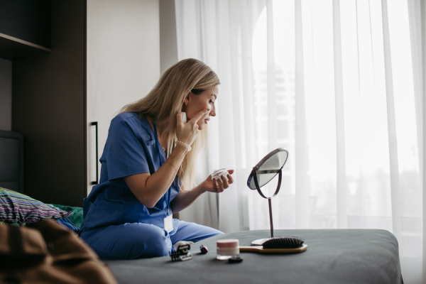 Nurse, doctor getting ready for work in the morning, applying moisturiser cream, doing makeup, skincare in bedroom. Work-life balance for healthcare worker.