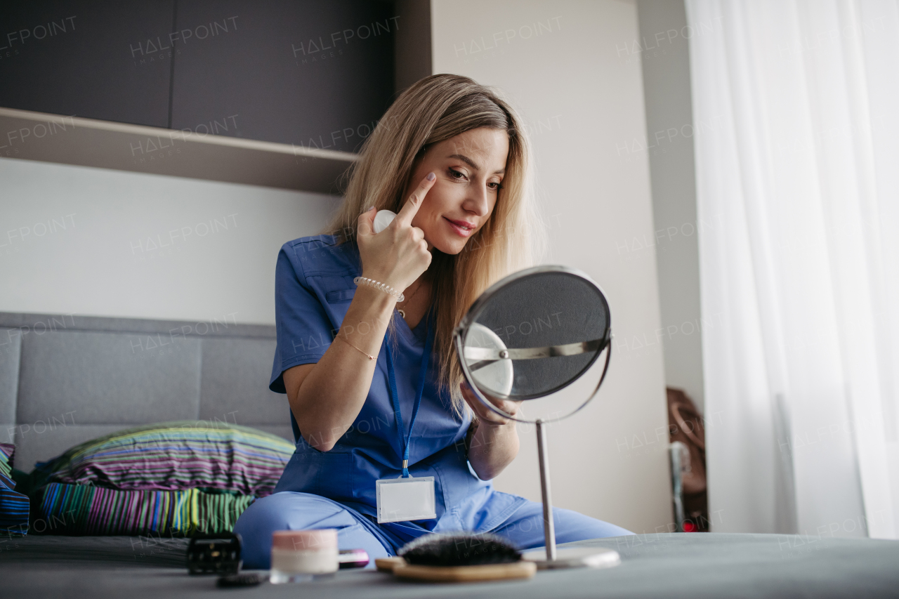 Female nurse or doctor getting ready for work in the morning, doing makeup, skincare in bedroom, dressed in scrubs. Work-life balance for healthcare worker.