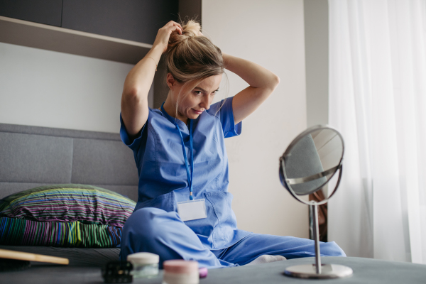 Female nurse or doctor getting ready for work in the morning, brushing hair, making ponytail in bedroom, dressed in scrubs. Work-life balance for healthcare worker.