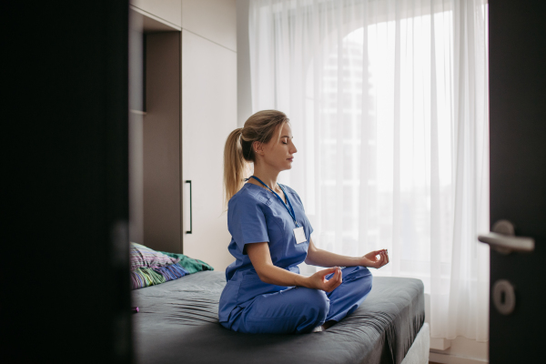 Female nurse or doctor enjoying free time at home after work, meditating in bedroom. Work-life balance for a healthcare worker.