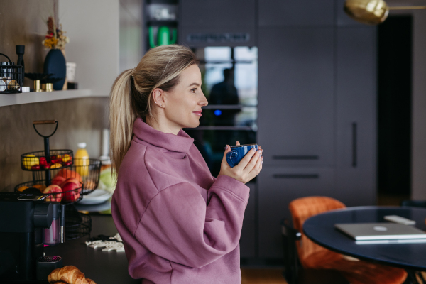 Side view of smiling woman enjoying cup of coffee at home after work, standing in kitchen wearing pink hoodie. Work-life balance for healthcare worker. Ear piercings.