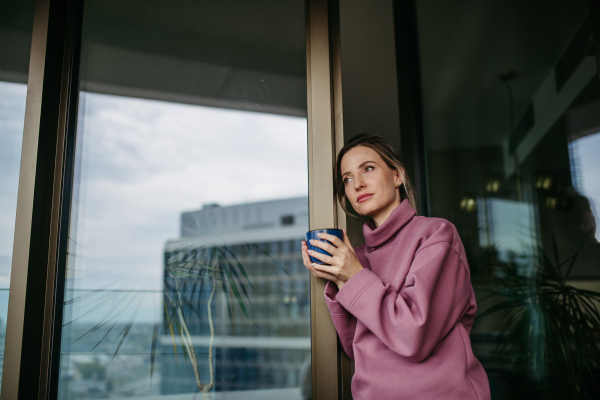 Female nurse enjoying cup of coffee at home after work, standing by window pink hoodie. Work-life balance for healthcare worker. Woman enjoying view from balcony.