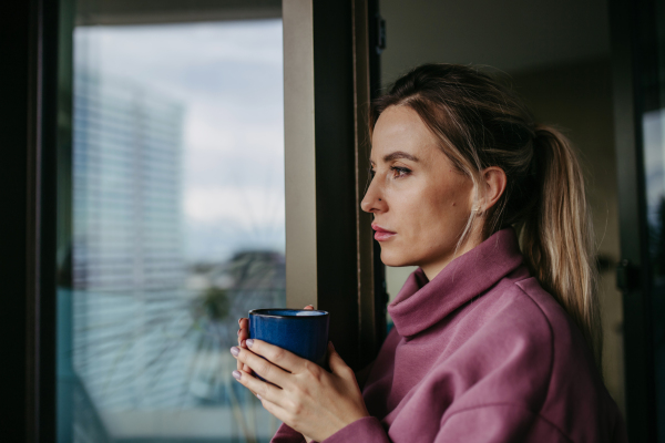 Woman enjoying cup of hot tea, coffee at home after work, standing by window wearing pink hoodie. Work-life balance for healthcare worker. Nose piercings.