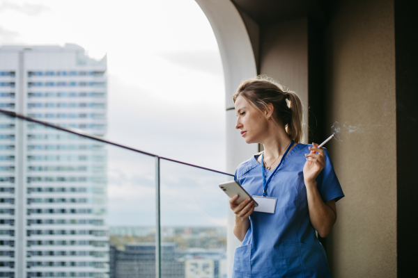 Female nurse smoking cigarette at balcony after work. Doctor feeling overwhelmed and exhausted at work, taking break and smoking.