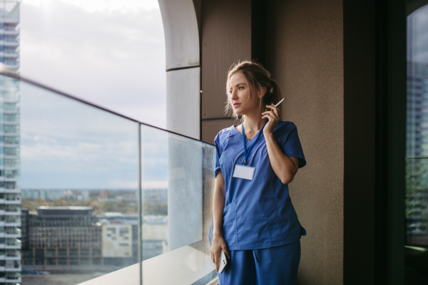 Female nurse smoking cigarette at balcony after work. Doctor feeling overwhelmed and exhausted at work, taking break and smoking.