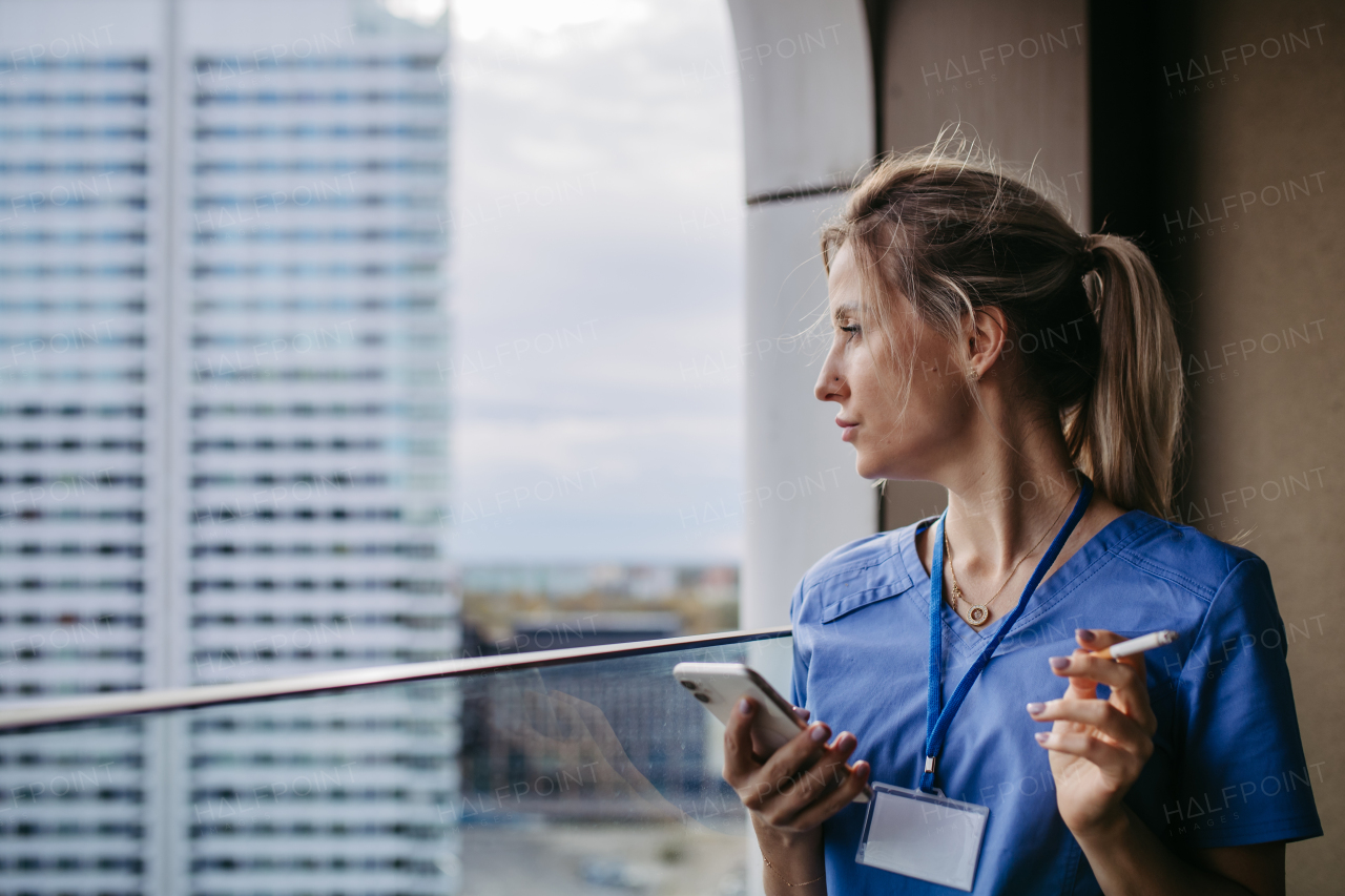 Nurse smoking cigarette at balcony after work, scrolling on smartphone. Doctor feeling overwhelmed at work, taking break. Stresfull at hospital, clinic.