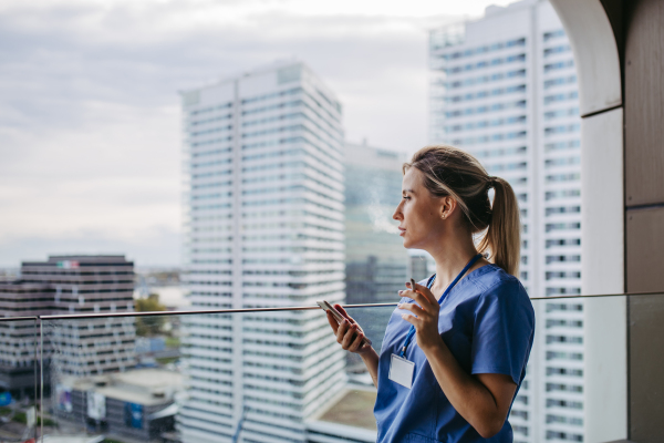 Nurse smoking cigarette at balcony after work, scrolling on smartphone. Doctor feeling overwhelmed at work, taking break. Stresfull at hospital, clinic.