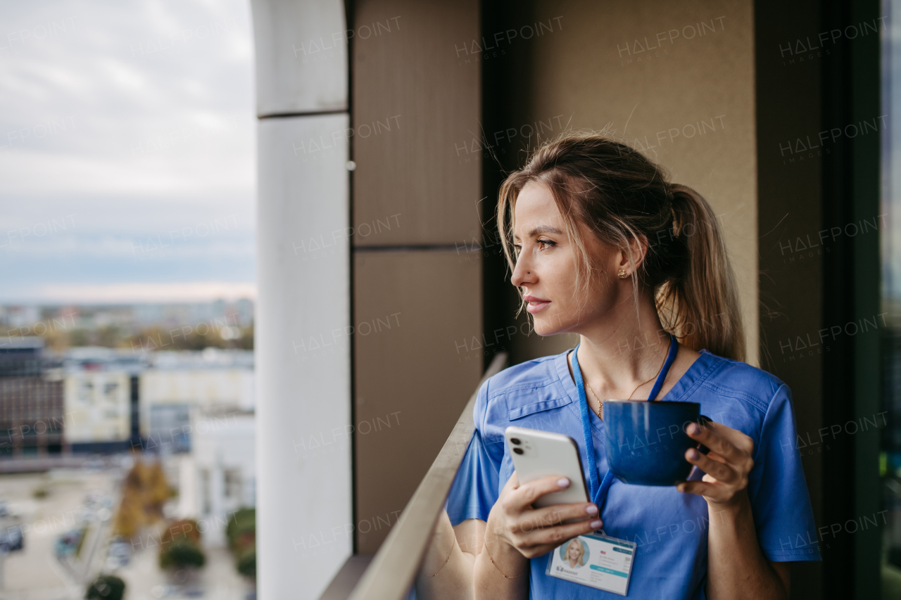 Female nurse enjoying cup of coffee at home after work, standing by window. Morning tea before work. Work-life balance for healthcare worker. Woman enjoying view from balcony.