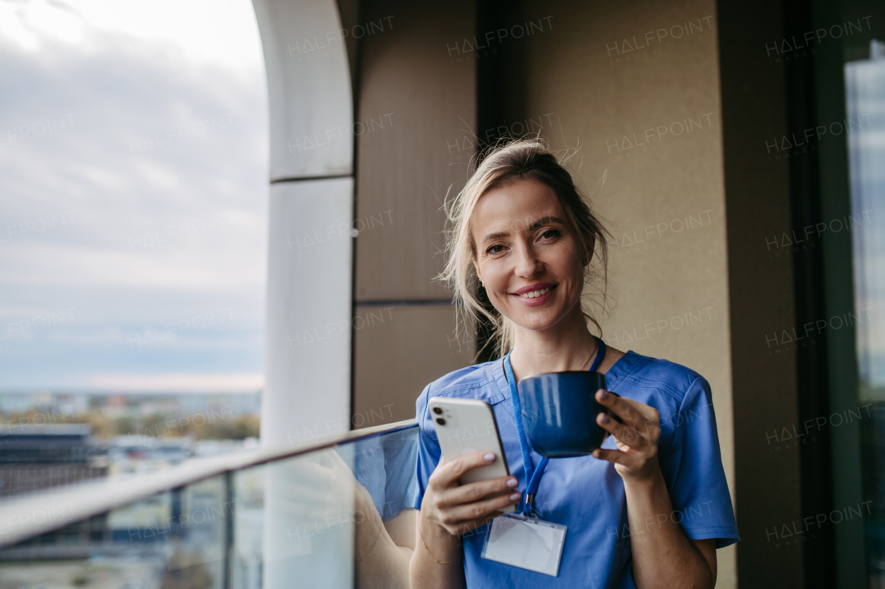 Nurse, doctor in uniform taking break, standing on hospital balcony and drinking coffee, scrolling on smartphone. Staff break area. Work-life balance of healthcare worker.