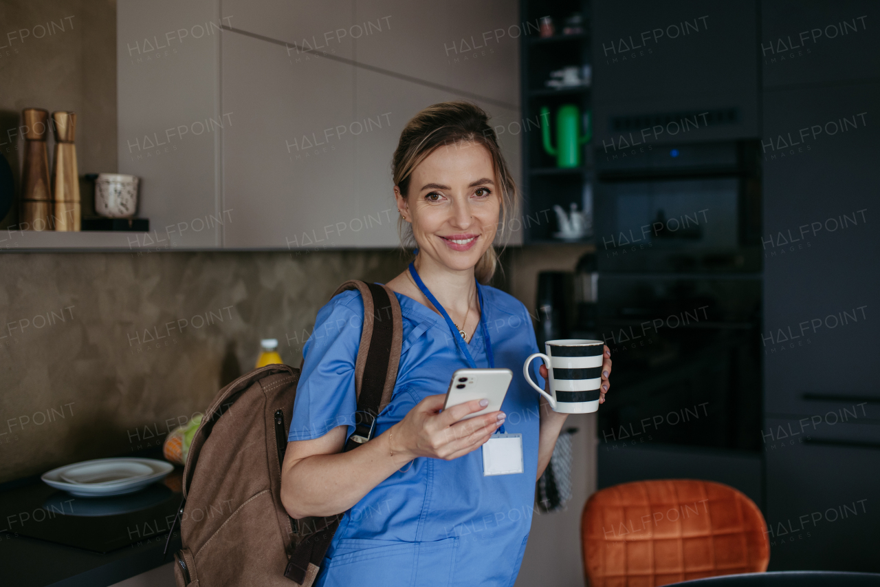 Female nurse, doctor getting ready for work, reading message on smartphone, drinking coffee, leaving house in scrubs with backpack. Work-life balance for healthcare worker.