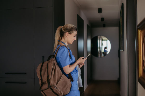 Female nurse, doctor getting ready for work, reading message on smartphone, leaving house in scrubs with backpack. Work-life balance for healthcare worker.