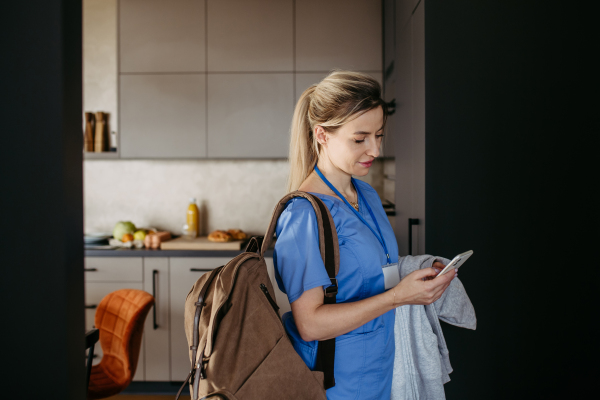 Female nurse, doctor getting ready for work, reading message on smartphone, leaving house in scrubs with backpack. Work-life balance for healthcare worker.