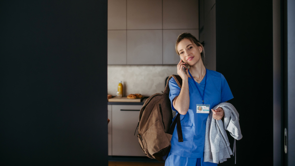 Female nurse, doctor getting ready for work, phone calling on smartphone, leaving house in scrubs with backpack. Work-life balance for healthcare worker.