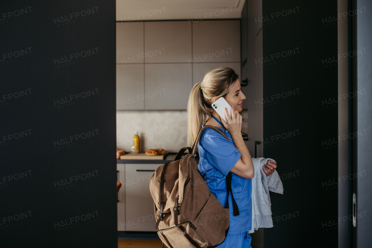 Female nurse, doctor getting ready for work, reading message on smartphone, leaving house in scrubs with backpack. Work-life balance for healthcare worker.