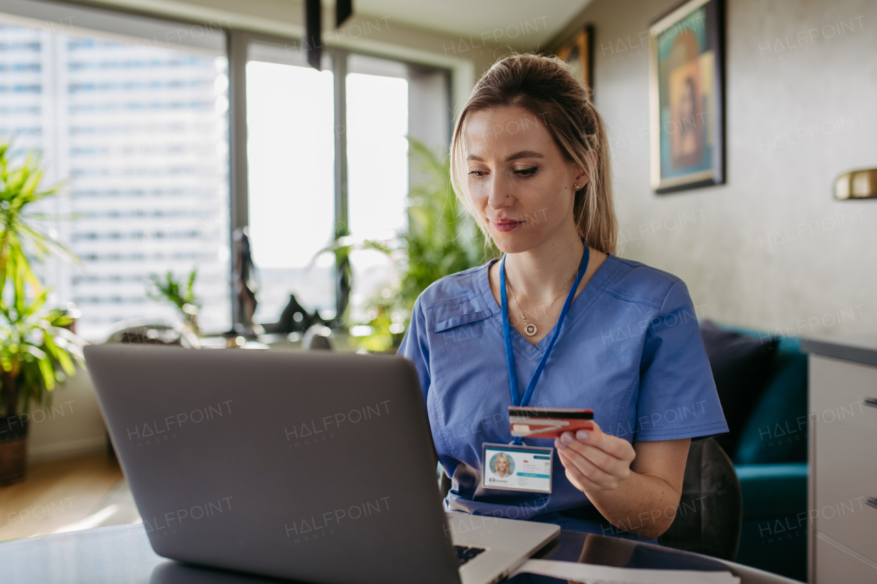 Nurse paying bills online with debit or credit card, working on notebook. Doctor in blue uniform shopping online, Work-life balance for healthcare worker.