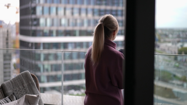 Female nurse enjoying cup of coffee at home after work, standing by window pink hoodie. Work-life balance for healthcare worker. Woman enjoying view from balcony.