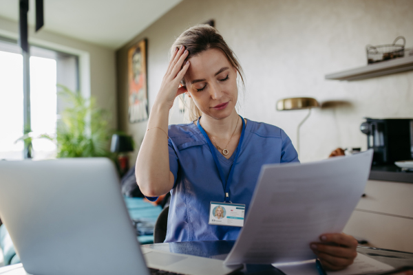 Nurse paying bills online, sitting in living room, working on notebook. Doctor in scrubs dealing with work issues from home, after workday, reading medical record, telemedicine consultation. Work-life balance for healthcare worker.