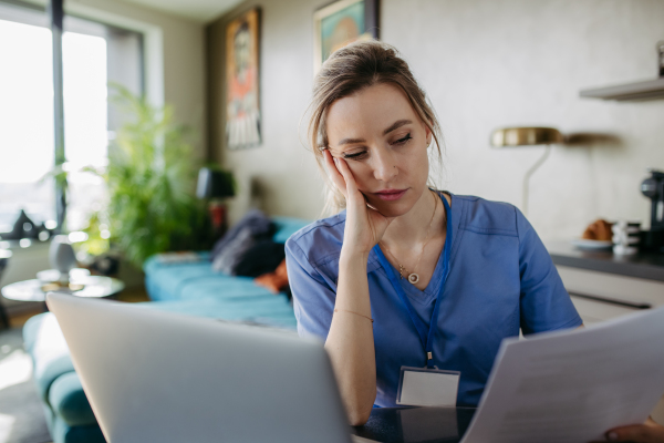 Nurse paying bills online, sitting in living room, working on notebook. Doctor in scrubs dealing with work issues from home, after workday, reading medical record, telemedicine consultation. Work-life balance for healthcare worker.