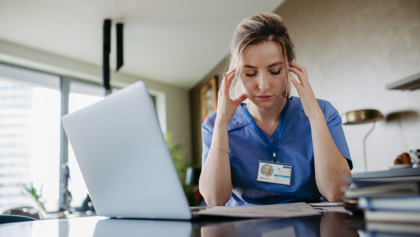 Nurse paying bills online, sitting in living room, working on notebook. Doctor in scrubs dealing with work issues from home, after workday, reading medical record, telemedicine consultation. Work-life balance for healthcare worker.