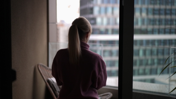 Female nurse enjoying cup of coffee at home after work, standing by window pink hoodie. Work-life balance for healthcare worker. Woman enjoying view from balcony.