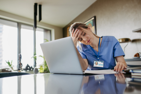 Nurse paying bills online, sitting in living room, working on notebook. Doctor in scrubs dealing with work issues from home, after workday, reading medical record, telemedicine consultation. Work-life balance for healthcare worker.